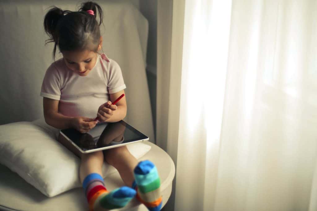 A young girl sits indoors using a tablet, enjoying a learning activity.